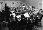 Bilden visar kungliga flottans musikkår år 1936, församlade i den då nya musiksalen på gamla Sparre. Här på fotot höjer den nya musikdirektören Harry Olsson (Brocktorp) sin taktpinne över en vältrimmad klarinettkår, vilken på sin tid var världsunik. Han kom från Hallands regemente och stannade här i Karlskrona till år 1944.