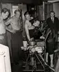 Översvämning på varuhuset Nordiska Kompaniet i Stockholm 1947. Fyra män, troligen brandmän, står runt en teknisk apparat med slangar och mätare. Mannen som sköter apparaten är klädd i uniformsjacka och mössa, hans byxor är uppkavlade. De övriga männen har bar överkropp, en av dem är tatuerad. I bakgrunden står en något äldre man i kavaj, skjorta och slips. Det är vatten på golvet.