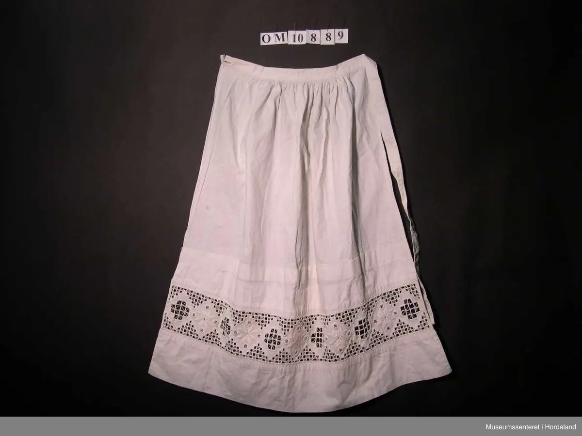 Form: bunadsforkle med linning og band i livet, knappa bak, innfeld broderi nedst på forkle
