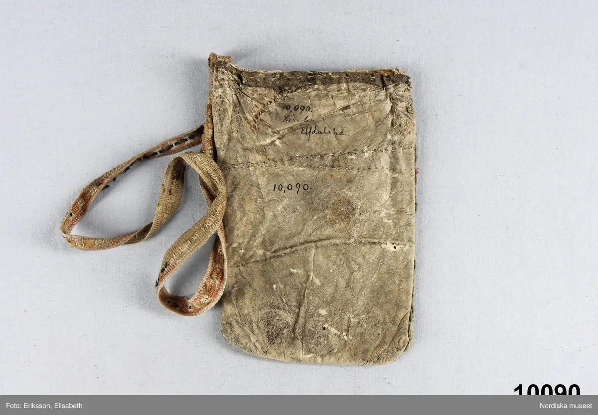 Rektangulär påse av vitt skinn med enkla blomfigurer samt initialer och årtal B F D 1790 av blå, röda och gröna vadmalsremsor applicerade med efterstygn av vitt lingarn,. Kanten prydd med bård av en blå och en röd yllerema trädda genom uppskurna slitsar i skinnet, i kanten fragment av gult ylle.
Mycket slitet linneband med upphämtamönster i gult, rosa och blått ullgarn fastsytt som axelband.
Insamlat av Nils Månsson Mandelgren vid mitten av 1800-talet.
/Berit Eldvik 2010-02-19