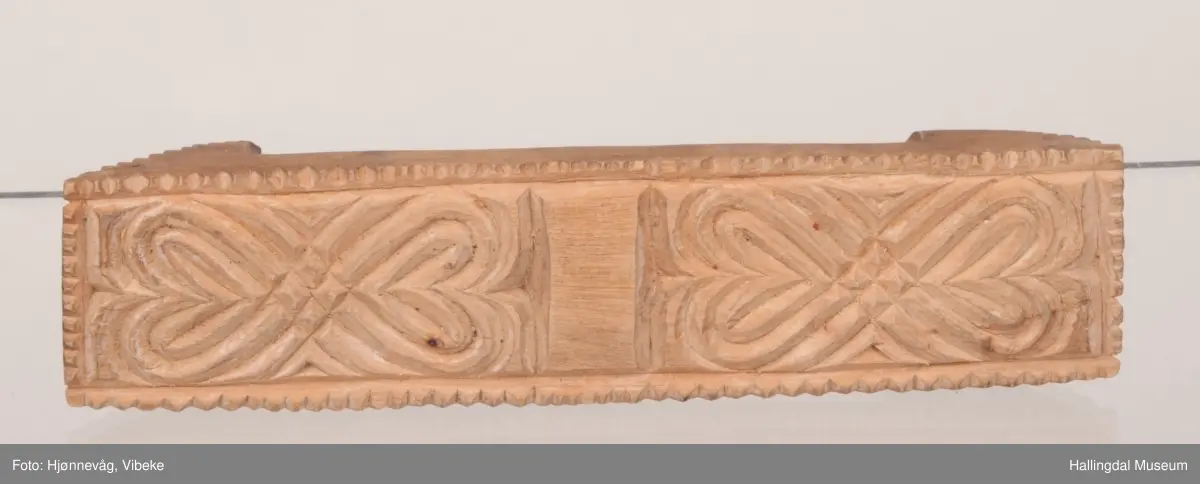 Rektangulær med skråttstilte flater i sidene. Mønsteret består i bunnen av to like felt, blomster dannet av to hjerter stilt med spissene mot hverandre. Tausnoingsborder langs kantene. Den ene sideflaten er nesten kvadratisk, hjertemønster omgitt av tausnoingsbord. Den andre flaten har en rett side nederst, oval øverst, hjertemønster og tausnoingsbord. Baksiden har håndtak, skåret i ett med stemplet. Buet med innskårne kryss på ryggen. Oppl. gml. prot.: smørknott.

23-7-1963. Å. Astrup.