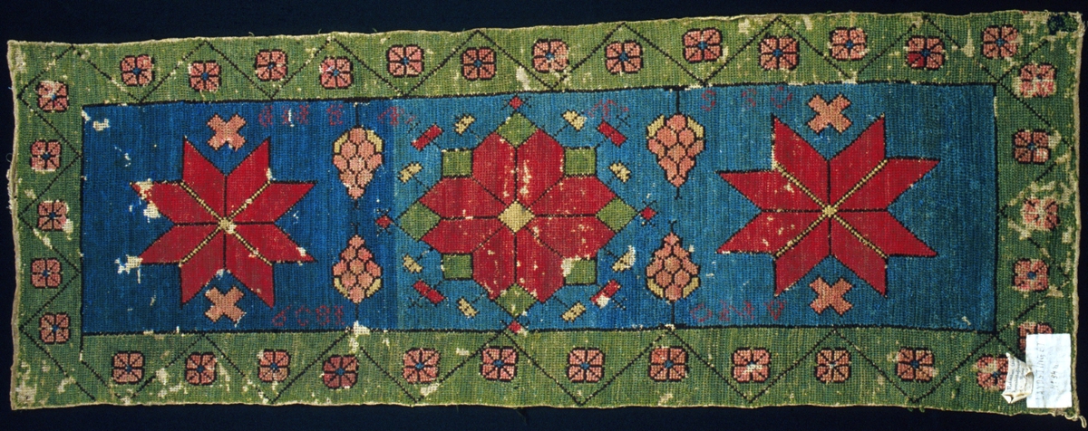 Dyna broderad i tvistsöm och korsstygn. Blått mittfält med en stjärnform i rött och grönt samt en spetsuddig stjärna i rött på var sida om denna. Kottliknande former och kryss i rosa som fyllnadsmönster. Runt om löper en 80-90 mm bred bård i grönt med rosa rosor och en mörkbrun sicksack-linje. Konturer i mörkbrunt. Broderad märkning i korsstygn i rött ullgarn: "ANO  1809" upptill och "OSS W W BMD" nedtill. Sydd på tvistbotten, 3-4 stygn/cm, med ullgarn; 2 eller 3 trådar tillsammans av s-spunnet garn eller 2 trådar tvinnade av en z-spunnen och en s-spunnen tråd. Broderiet är utfört övervägande i tvistsöm, konturer och mindre mönsterformer är sydda i korsstygn. Tekniken är tvåradig lodrät tvistsöm, man syr med stygnen ifrån sig, dvs. nedifrån och uppåt. Man startar alltid med det första stygnet i vänster hörn. Infållade kanter i kortsidorna, stadkanter i långsidorna. Tre stycken trasiga trådöglor på baksidan längs ena kortsidan för upphängning. Märkt på framsidan med påsydd tyglapp med texten: "LANDSTINGET N° 54 a". Dessutom en vidhängande pappersetikett med texten: "Malmöhus läns Hemslöjdsförening Malmö  Rönnebergs h:d".