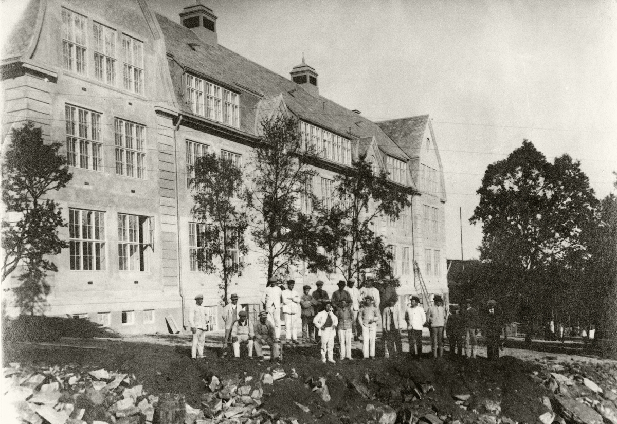 Fra byggingen av Harstad Folkeskole. Her står anleggsarbeidere, og muligens noen lærere eller medlemmer av kommunestyret(?), foran "byskolen", som sto ferdig i 1918.