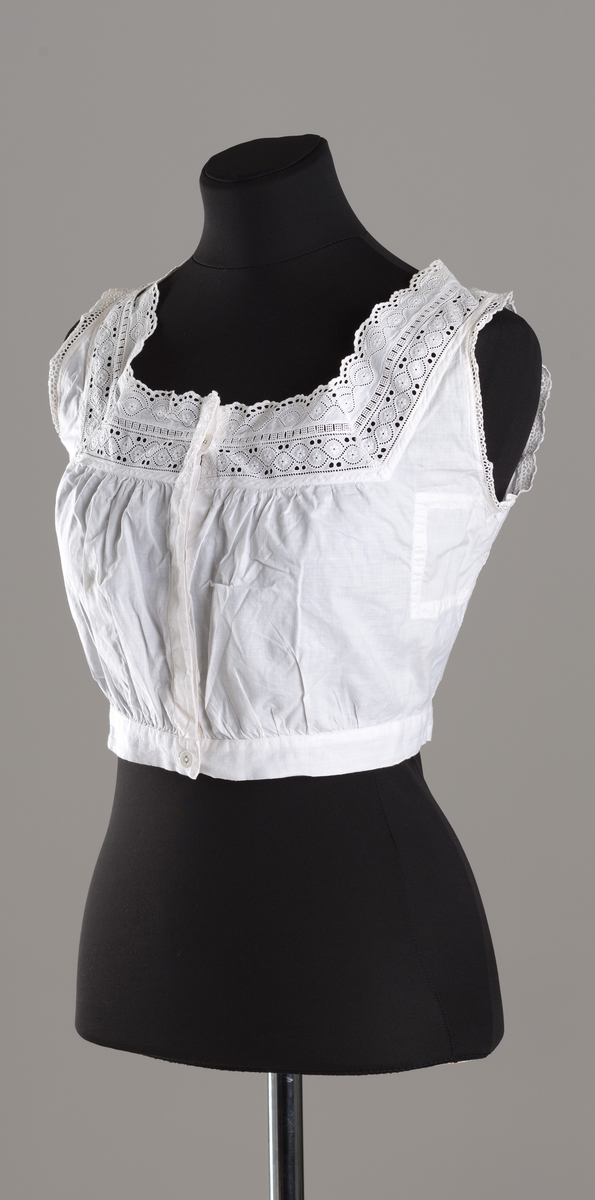 Hvitt underliv i tynt bommullslerret. Rynker over påsydd linning og på bryst. Firkantet hals dekorert med brede blonder foran og smalere blonder bak. Ermåpning kantet med smale blonder. Knappestolpe med skjult knapping, 4 knapper med hull, derav den nederste synlig. Knappene er trukket med hvitt lerret.