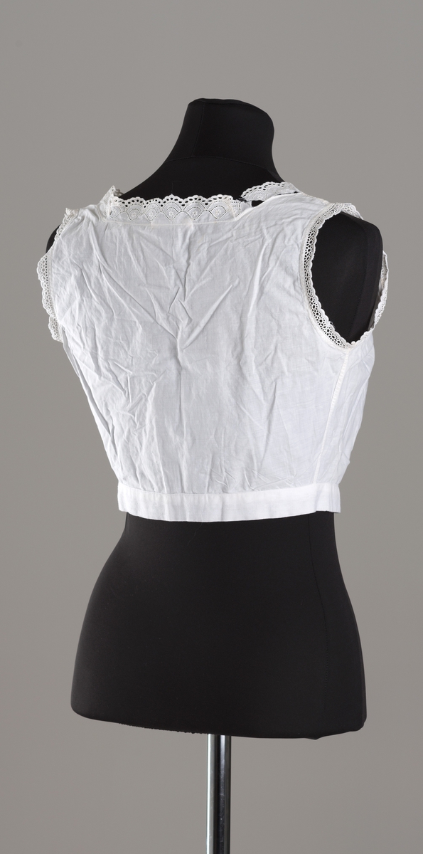 Hvitt underliv i tynt bommullslerret. Rynker over påsydd linning og på bryst. Firkantet hals dekorert med brede blonder foran og smalere blonder bak. Ermåpning kantet med smale blonder. Knappestolpe med skjult knapping, 4 knapper med hull, derav den nederste synlig. Knappene er trukket med hvitt lerret.