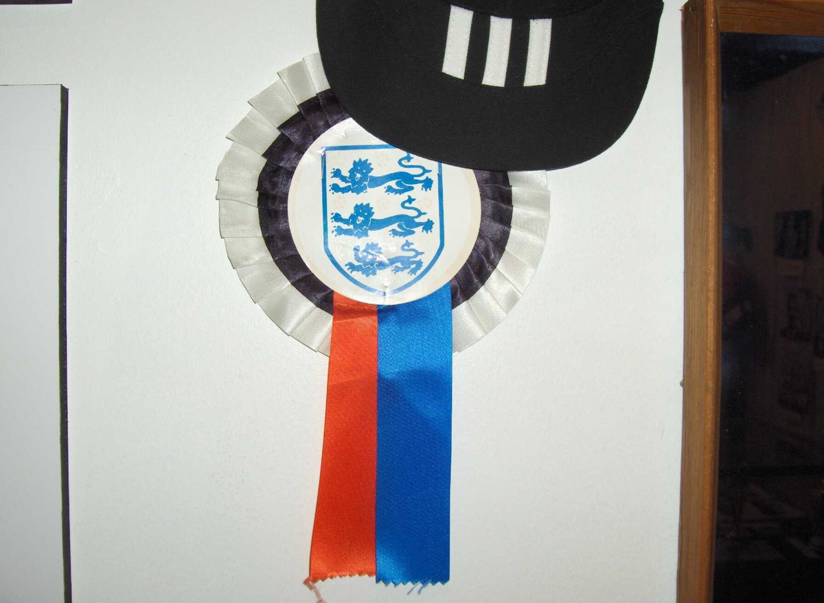 Pappsløyfe til å ha på brystet. Supporterutstyr England til EM i 1996, som gikk i England. De tre løvene som ble sunget om i sangen "Football's coming home" på denne tida.