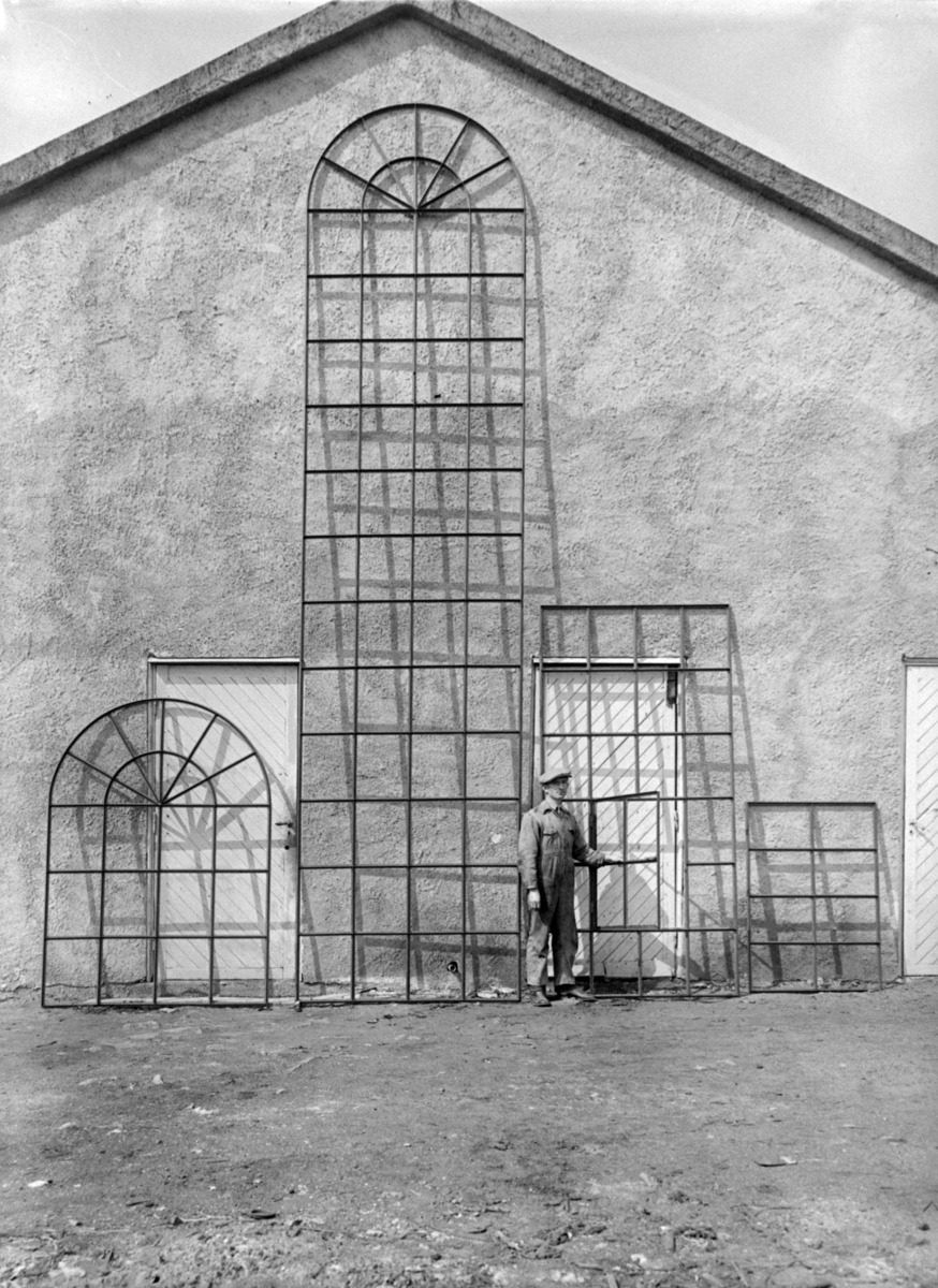 Metallfatninger til vindusglass stilt opp mot husvegg, og arbeider. Fredrikstad Jern og Metallindustri.