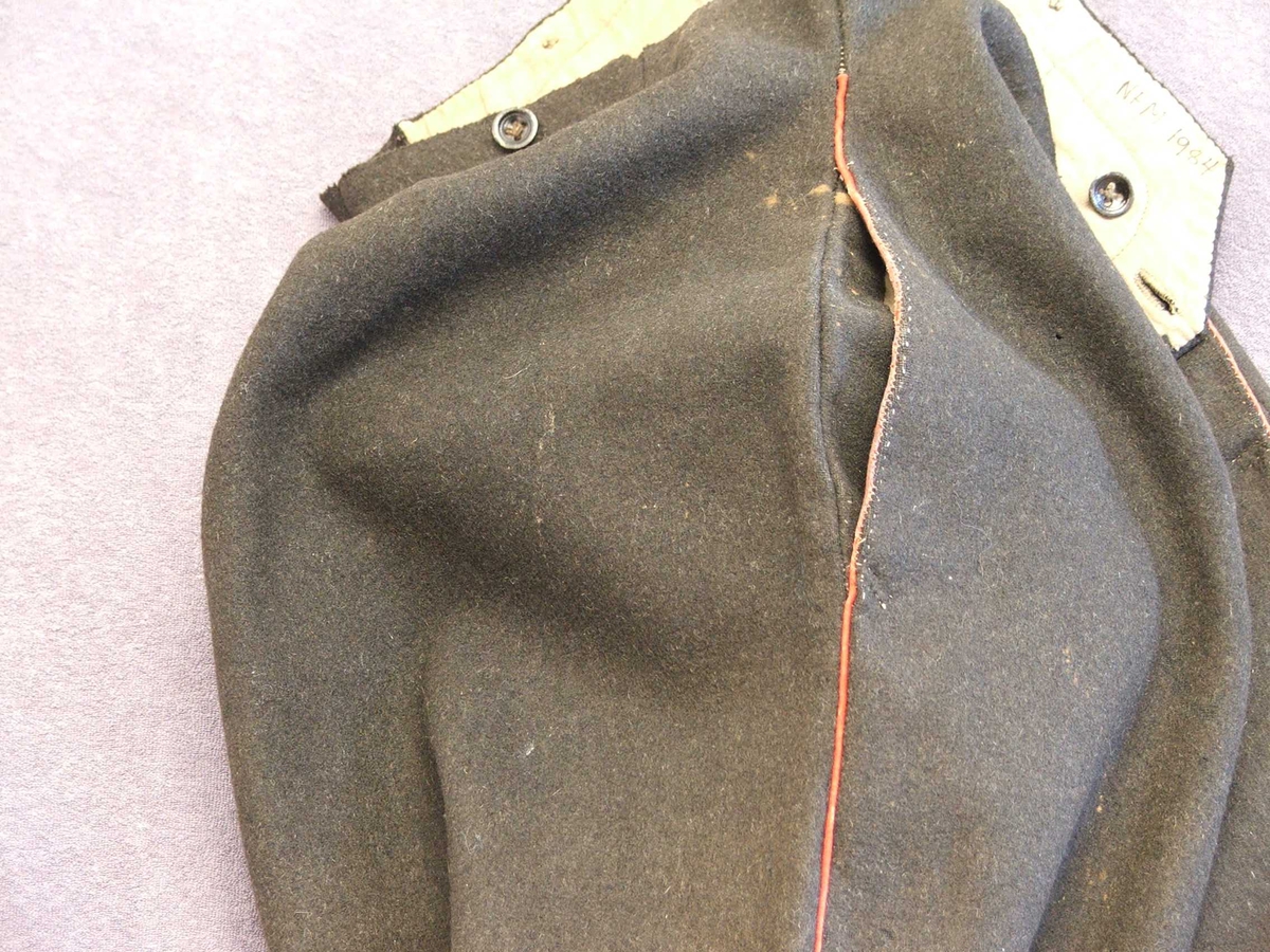 Uniformsbukse til mann. Svart med raud isydd stripe i sidesaumane. Fôra øvst ved lininga. Opning midt framme med fem knapphol. Ein knapp på kvar side av midten framme og bak. Kile isydd midt bak med eit lite hol på kvar side. Eit legg på kvar side av midtsaumen bak. Bak er påsydd to hemper i same stoff som buksa, hempene kan knappast saman. Metallknappar. Fire kilar isydd i skrittet.