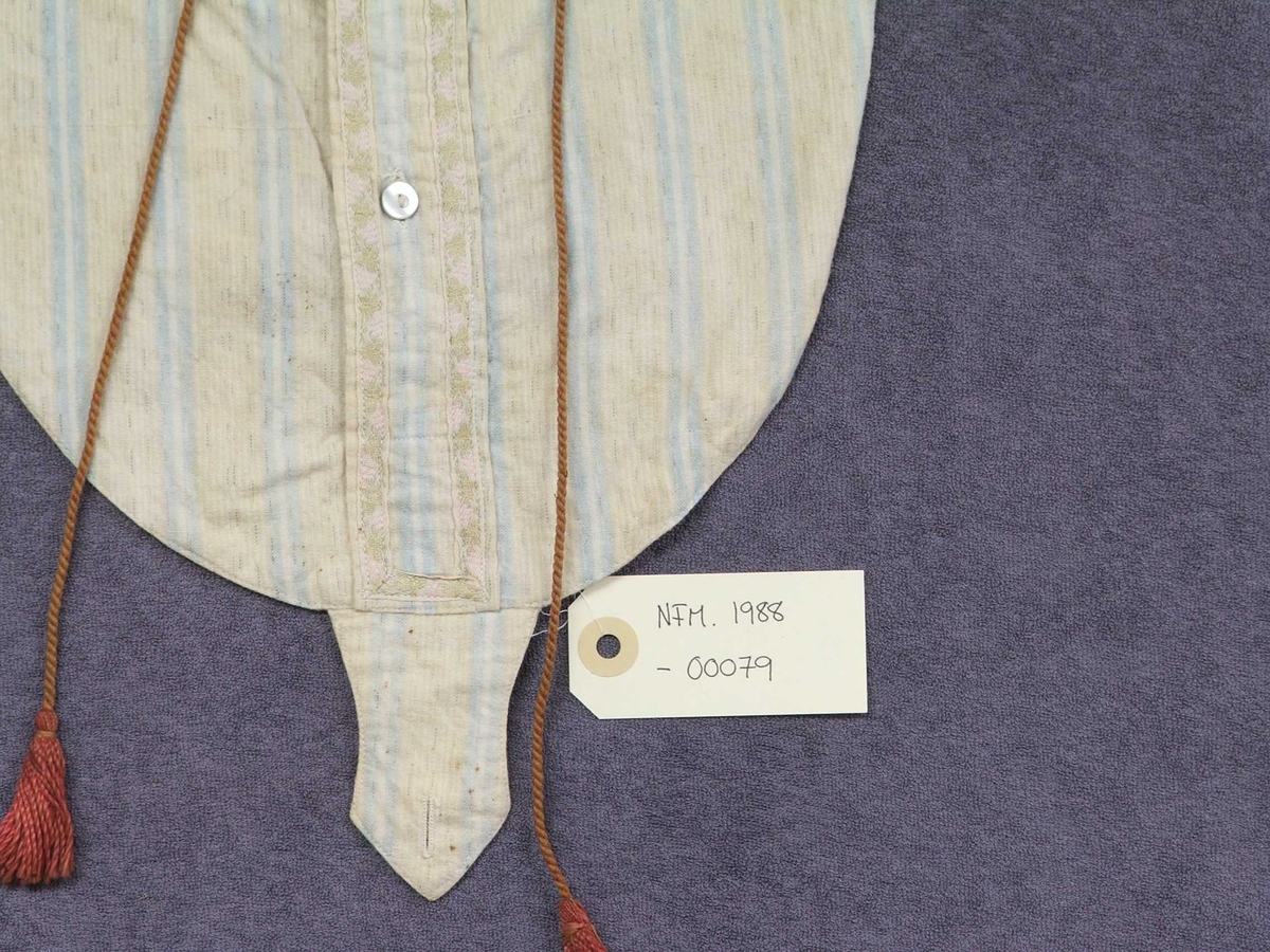 Skjortebryst. Oval form med knapping framme, opning til halsen, snippkrage med knappar ned på stoffet under. Nedst midt framme er sydd på ei "tunge" med knapphol i same stoff. Stoffet har lyseblå og kvite striper. Fôra med einsfarga stoff. Pynteband på krage og knappestolper med grønt og rosa. Brunraud snor m/duskar rundt halsen og heng ned framme.