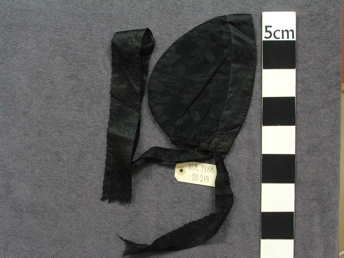 Svart "hue" til kvinne. Konehue. Typen er tett inntil hovudet. Med 4 cm kant fremst i glatt stoff, og med resten av utsida i svart stoff med innvove blomemønster.Lange band bak i nakken til knyting. Bak i nakken ein om lag 11 cm høg splitt, øvst i splitten er bandet trædd inn og i to hol på kvar side. Det er altså eitt band som kan snurpast ihop her. Fôra med lys gult og kvitt bomullsstoff.