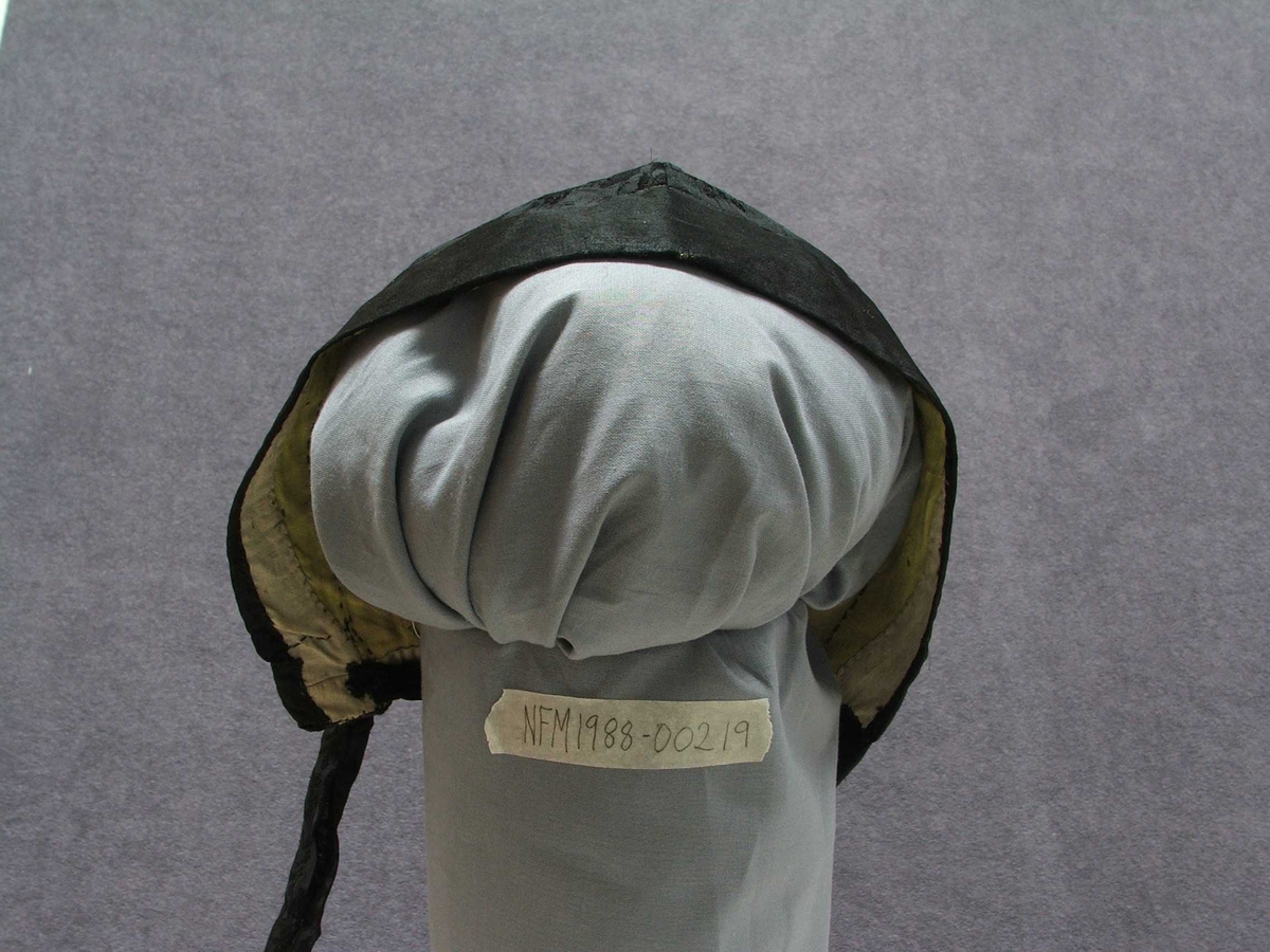 Svart "hue" til kvinne. Konehue. Typen er tett inntil hovudet. Med 4 cm kant fremst i glatt stoff, og med resten av utsida i svart stoff med innvove blomemønster.Lange band bak i nakken til knyting. Bak i nakken ein om lag 11 cm høg splitt, øvst i splitten er bandet trædd inn og i to hol på kvar side. Det er altså eitt band som kan snurpast ihop her. Fôra med lys gult og kvitt bomullsstoff.