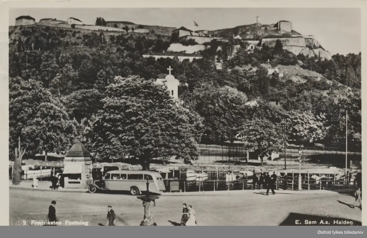 Halden - Wiels plass med Fredriksten festning og Immanuelkirken i bak- grunnen,  aviskiosk og en buss ses ved kanten av Tista.