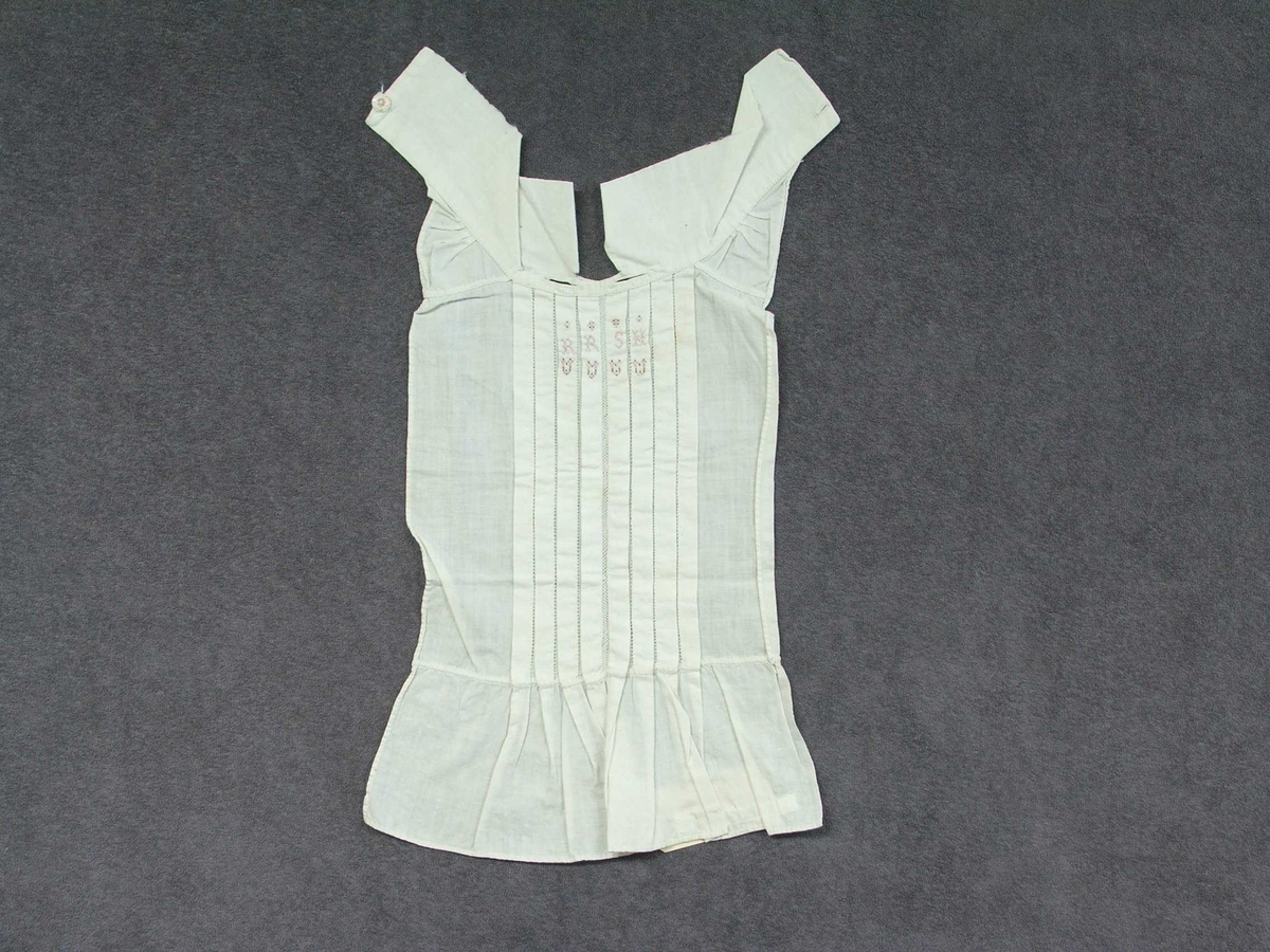 Skjortebryst til å ha framme på brystet. Til mann. Forseggjort saum. Krage til å knappe bak med knapp og knapphol. Eit knapphol på kvar side av kragen. Fire pyntefaldar på kvar side av midten framme på brystet. Det er ein slags netting langs midten og holsaum mellom kvar fald. Sydd på omlag 10 cm kant nedst utan pynt. Brodert fire små hjarte i rosa og brune krossting under fire bokstavar og ståande firkantar over. Alt er kanta.