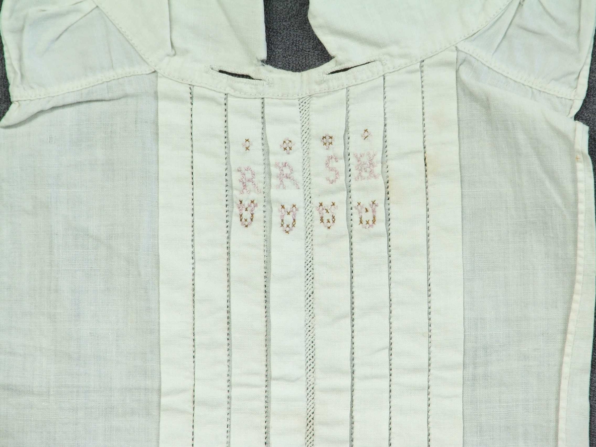 Skjortebryst til å ha framme på brystet. Til mann. Forseggjort saum. Krage til å knappe bak med knapp og knapphol. Eit knapphol på kvar side av kragen. Fire pyntefaldar på kvar side av midten framme på brystet. Det er ein slags netting langs midten og holsaum mellom kvar fald. Sydd på omlag 10 cm kant nedst utan pynt. Brodert fire små hjarte i rosa og brune krossting under fire bokstavar og ståande firkantar over. Alt er kanta.