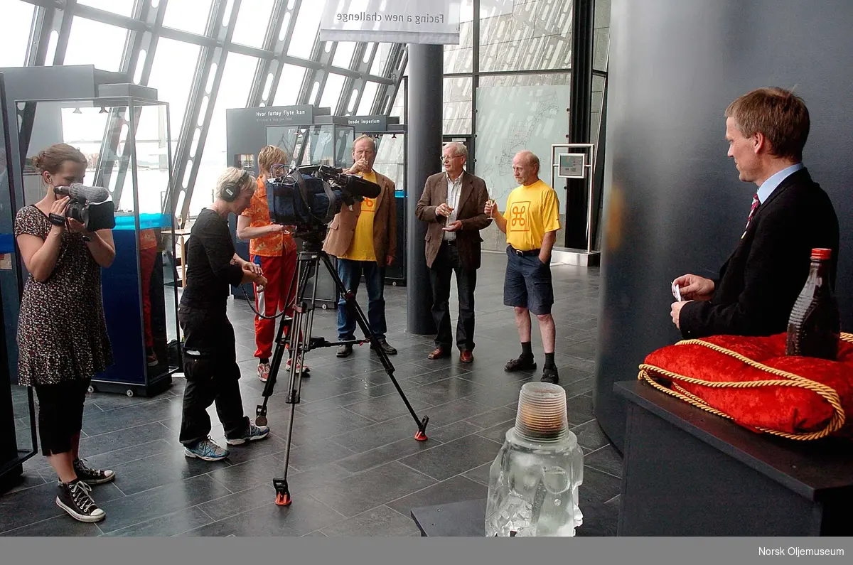 Flaske med "Den første olje" -  råolje fra det som skulle bli Ekofiskfeltet, skal stilles ut på Norsk Oljemuseum i Stavanger.
Anledningen er TV-opptak til NRK-programmet "Norsk Attraksjon" som ble vist på TV i september 2009.         
Som seg hør og bør skal en utstillingsåpning markeres på behørig vis.
Direktør Finn E. Krogh ved Norsk Oljemuseum skal holde åpningstale, mens NRK-fotograf Kristin Helgeland Hauge er i aksjon bak kamera.
Assistent Ingvil Teige Stiegler t.v. filmer også opptrinnet, mens de andre involverte bivåner det hele.
I forgrunnen står en borekrone i krystall med oljeinnhold fra det aller første oljefunnet til Esso på norsk sokkel, men feltet var ikke drivverdig, så glassflasken med råolje på puten t.h. i bildet markerer "Den første olje" ettersom feltet var til de grader drivverdig, og til dags dato har produsert olje i over 40 år. 