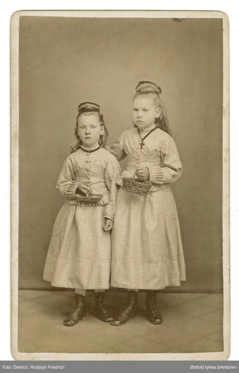 To barn, jenter, i finkjole med kurv og oppsatt hår, knappe støvler. 
Ukjente personer, ant. Fredrikstad eller Sarpsborg..

Fotografens gummistempel på revers (bakside) antyder at det er et av de tidligste fotografiene til Dietrich, kanskje like etter 1865, da han startet atelieret i Fredrikstad.
