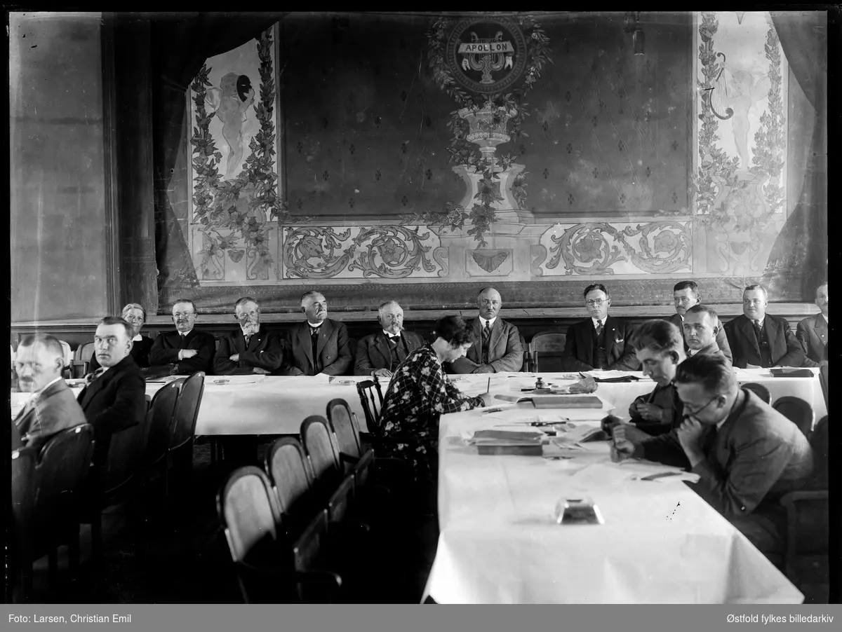 Østfoldutstillingen i Sarpsborg 1930.  
Komitemøte, interiør fra teatersal. scene med sceneteppe. Apollon  (lyre), menn, kvinne skriver. Ukjent sted, Sarpsborg? Klappstoler, Festiviteten?
Vandrelærer Ole Kjenner i midten, nr. 5 fra venstre (biavlsnæring).