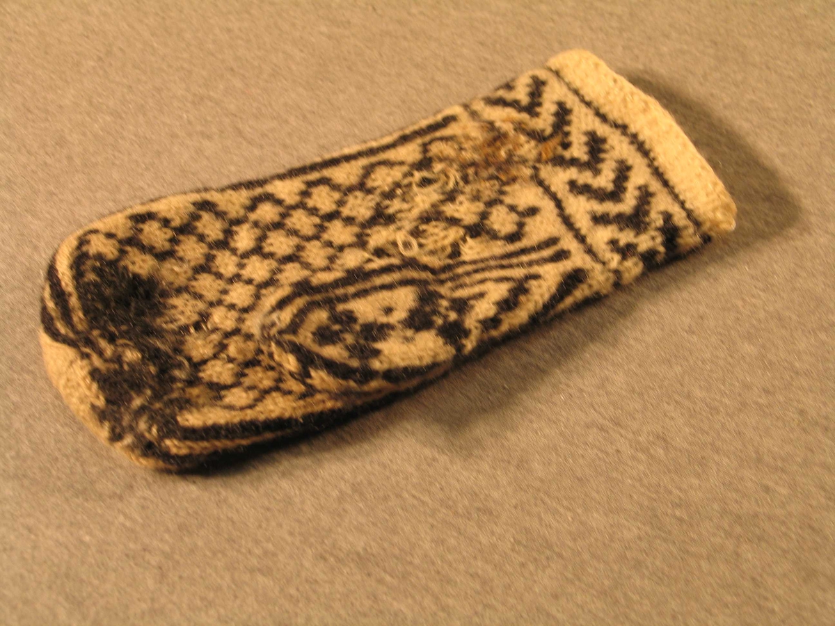 Handstrikka venstre-vott i såkalla selbumønster. Brot med to rette og ein rang over 2 omgangar, eit felt med liggande v-ar og sjølve votten med to store åttebladroser.