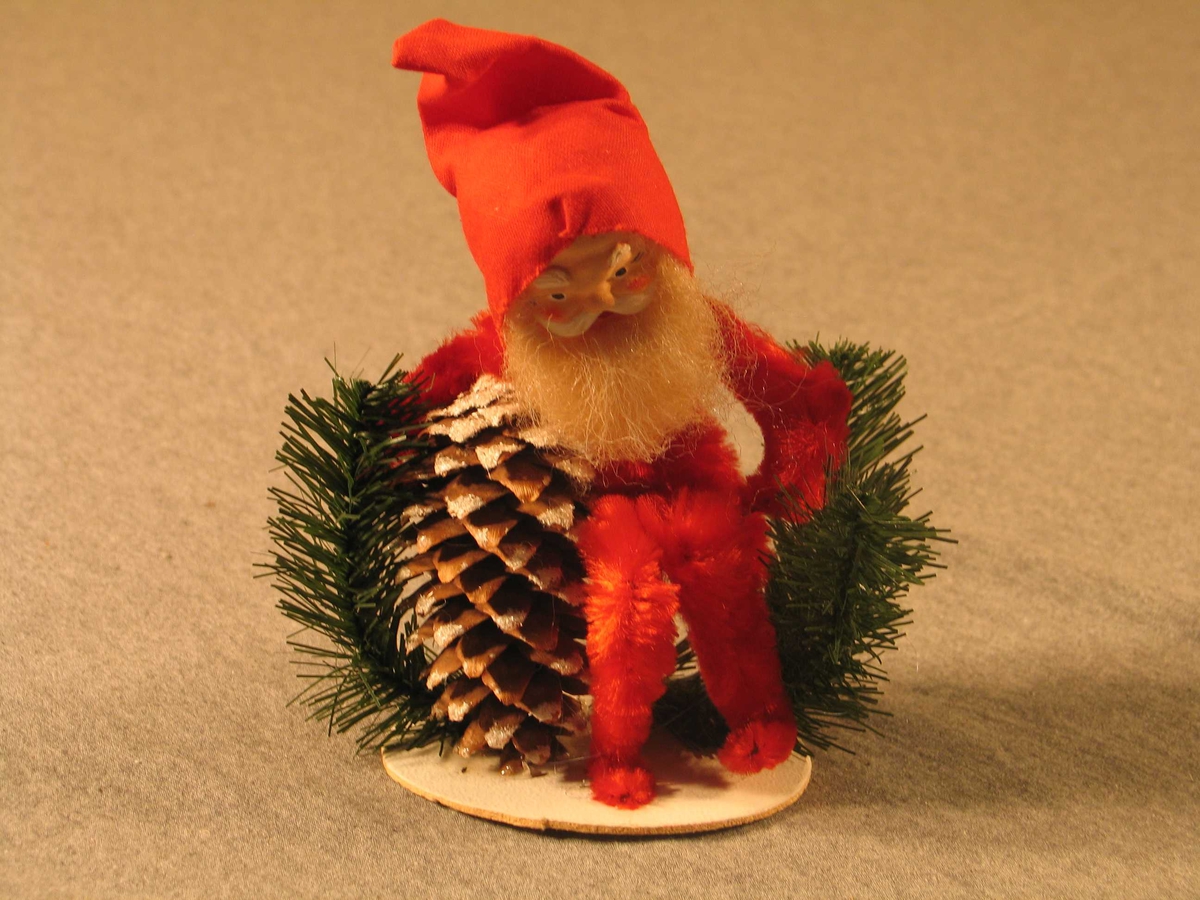 Julenisse av piperensar, i sittande stilling utan å ha nko å sitte på. Nissen er laga av piperensar med hovud av pappmache. Måla ansikt, skjegg og nissehue. Nissen held rundt ei krankongle. Plastimitasjonar av granbar.
Figuren er limt på ei papplate.