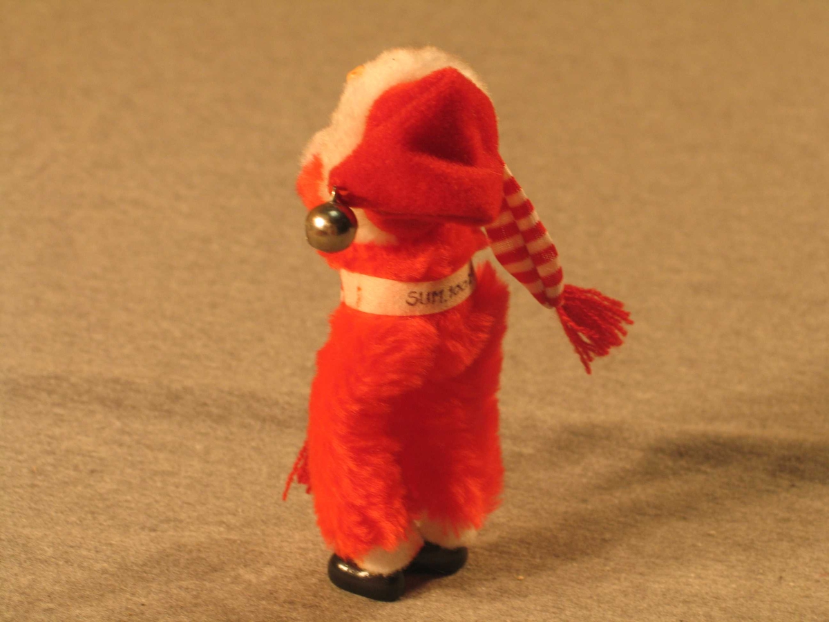 Nisse i heil flossdrakt. Kvitt belte og kantingar. Ansikt av plast. Bjelle i toppen av hua. Nissen held på eit langt sjal i raude og kvite striper.