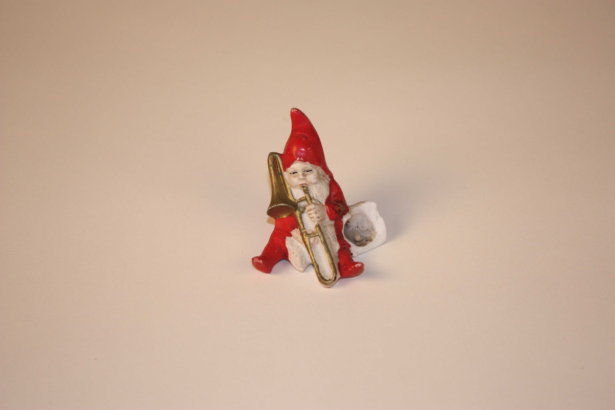 Nisse som sit og spelar trekkbasun. Ved sida av har han ei bøtte som det kan settast juletrelys i. Nissen har langt skjegg og hue med spiss.