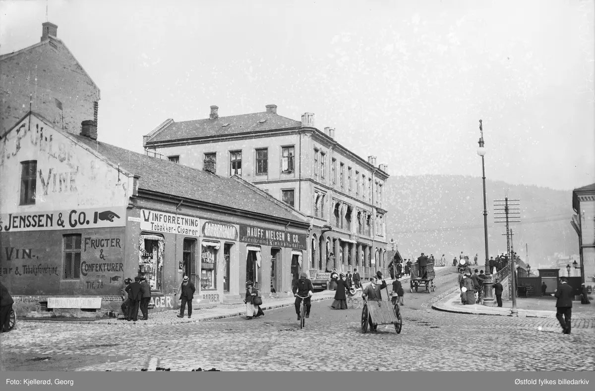 Fra Strømsø torg mot gamle bybrua i Drammen.  Selv om negativ eske er merket 1910 er bildet fra tidligst sommer 1909. Ingen ledninger til trikken.
Reklame for 
Bjørnstad Læderhandel.
Hauff. Nielsen & Co, Isenkram, kjøkkenutstyr.
Barbersalon.
Jenssen & Co, Vinforretning, Tobakker og Cigarer. Frukter og Confekturer.
Apothek.