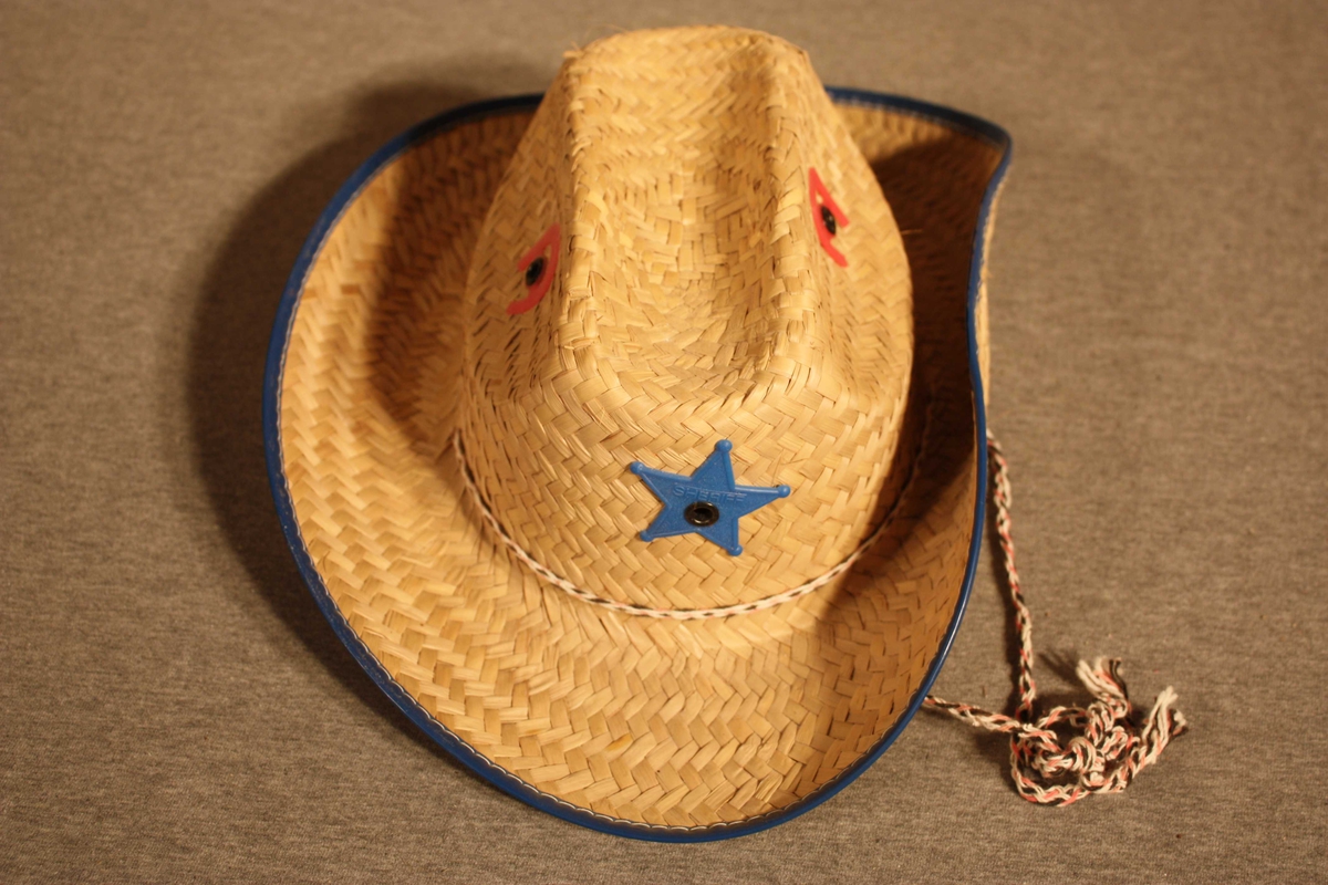 Stråhatt med påsydd blå plastlist rundt bremmen og plaststjerne og plasthestesko festa på pullen. Bomullstau som hakeband