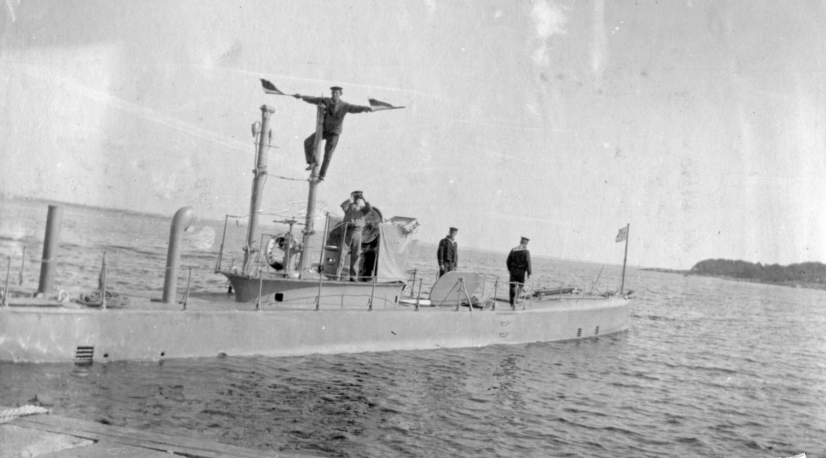 Karlskrona 1914 - Man står på ubåt och signalera med flaggor i händerna. Tre andra matroser står i bakrunden på ubåtsdäck.