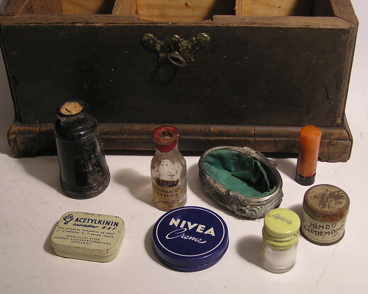 Rektangulær skrin med lokk og nøkkelhull. Liten nøkkel ligger i hullet.  Fotliste. Rommet er inndelt og inneholder små medisinflasker, krem og hodetabletter.
Ligner liten skipskiste.