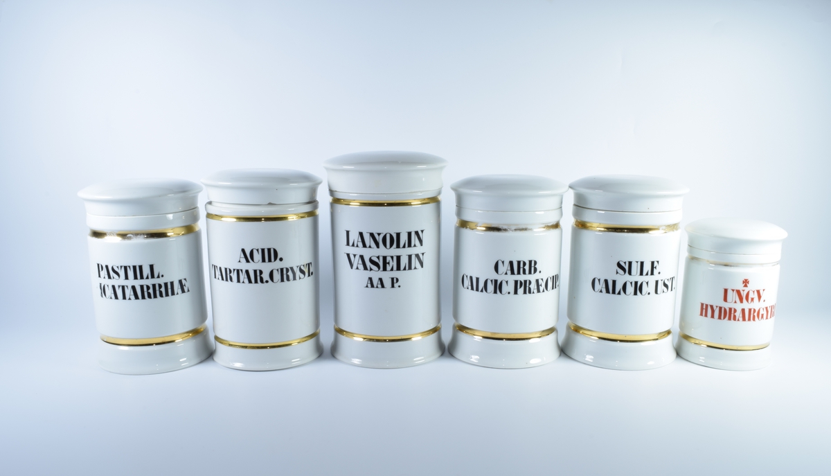 Seks sylindriske porselenskrukker med flatt lokk. Samtlige er dekorert med gullborder øverst og nederst og har skrift i svart eller rødt.