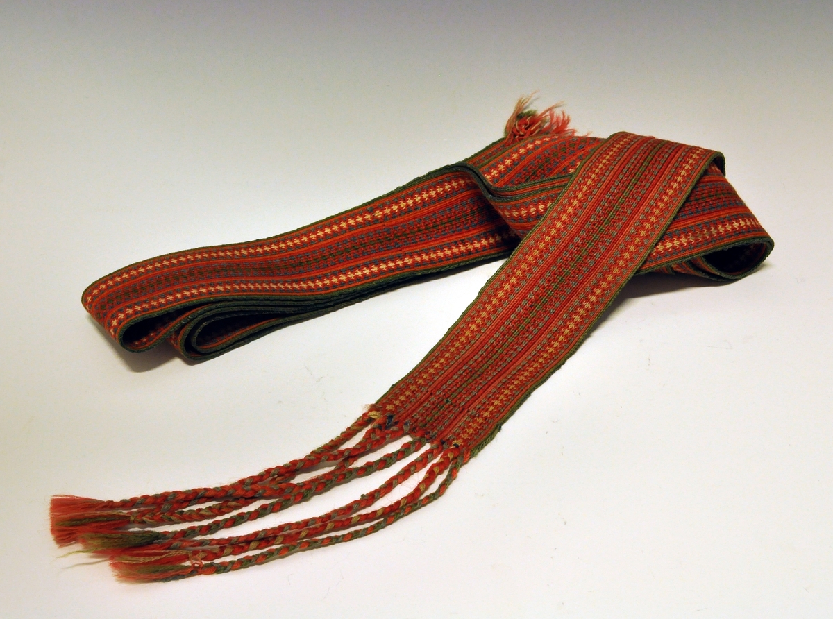 Belte til folkedrakt, brikkevevet. Frynser i den ene enden, de er flettet. Den andre enden er det renning uten avslutning.