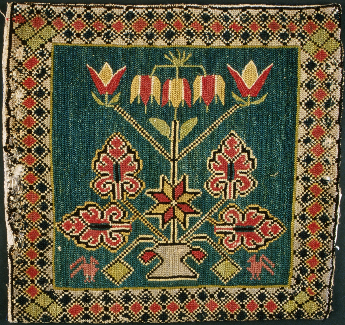 Jynne broderad i tvistsöm. Kejsarkrona på hög stängel mellan fyra akantusblad samt en spetsuddig stjärna med åtta vingfält. Buketten växer upp ur en urna. Två fåglar nedtill, diagonalmönstrad rutbård runt om. Grön botten, mönster i rött, gult, blått, rosa, ljusgrönt, mörkblått, grått, svart och naturvitt. Broderiet är utfört i traditionell bunden tvistsöm men också i en mer fri typ av tvistsöm. Runt vissa mönsterformer har konturer sytts, liknande de konturer som läggs i flamskvävning. Enstaka stygn, framför allt de svarta och naturvita konturstygnen, har sytts med korsstygn. Sydd på panamaväv i linne, 14 trådar/cm, med tvåtrådigt ullgarn och två trådar tillsammans av entrådigt ullgarn. Jynnet är öppet längs en sida. Baksida i blått satinvävt halvylletyg av yngre datum. Varp i två nyanser blått tvåtrådigt bomullsgarn, inslag i två nyanser blått entrådigt ullgarn, två trådar tillsammans. Lagad på några ställen. Märkt med påsydd tyglapp med texten: "Tillhör Malmöhus läns Hemslöjdsförening". Dessutom märkt med ett påsytt tygband med texten: "Sytt av Bolla Jönsdotter Östarp dotter till Bengta Persd. från Gästg. gården i Everlöv, g. m Helge Nilsson f. 1794 + 1784 g. 1814".