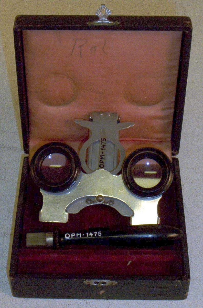 Instrument i forniklet stål, to linser, stillbar i høyde, forsynt med et sort håndtak til å sette inn i et spor. Instrumentet oppbevares i et sort etui. På innsiden av lokket merket "Rol".