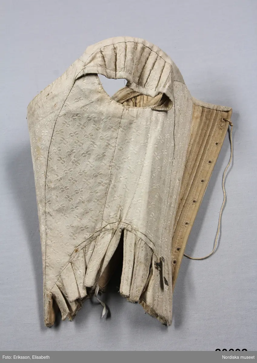 Huvudliggaren:
"Snörlif. Af hvitt siden, från 1770-talet."

Liv till hovdräkt s.k. "robe á la cour" av ursprungligen vit sidendroguet. Tyget förmodligen svensk tillverkning (Myrman?) se 1700-tals textil, A. Berchs samling, sid 171. Yttertyget är skuret i nio delar med mittstycke fram som löper ned i snibb samt fyra delar å varje sida med snörning mitt bak. Grundlivet tillverkat av oblekt linnelärft är helt styvat med spröt av valben trädda i sydda vertikala kanaler samt med en skena av järn som löper runt urringningen och planschett mitt fram. Grundlivet troligen klistrat med papper för att hålla en viss form innan yttertyget monterats på. Ärmholkar bestående av å varje sida tio hopsydda delar som lagts i veck. Tillskärningen följer helt den gängse för robe de cour. Se Waugh, Nora, The Cut of Womens Clothes, s. Längs sidorna i midjehöjd en tunn snodd avsedd att haka fast släpet i. Livet ursprungligen buret med kjol och släp i samma tyg.
/Ingrid Roos 2007-03-06