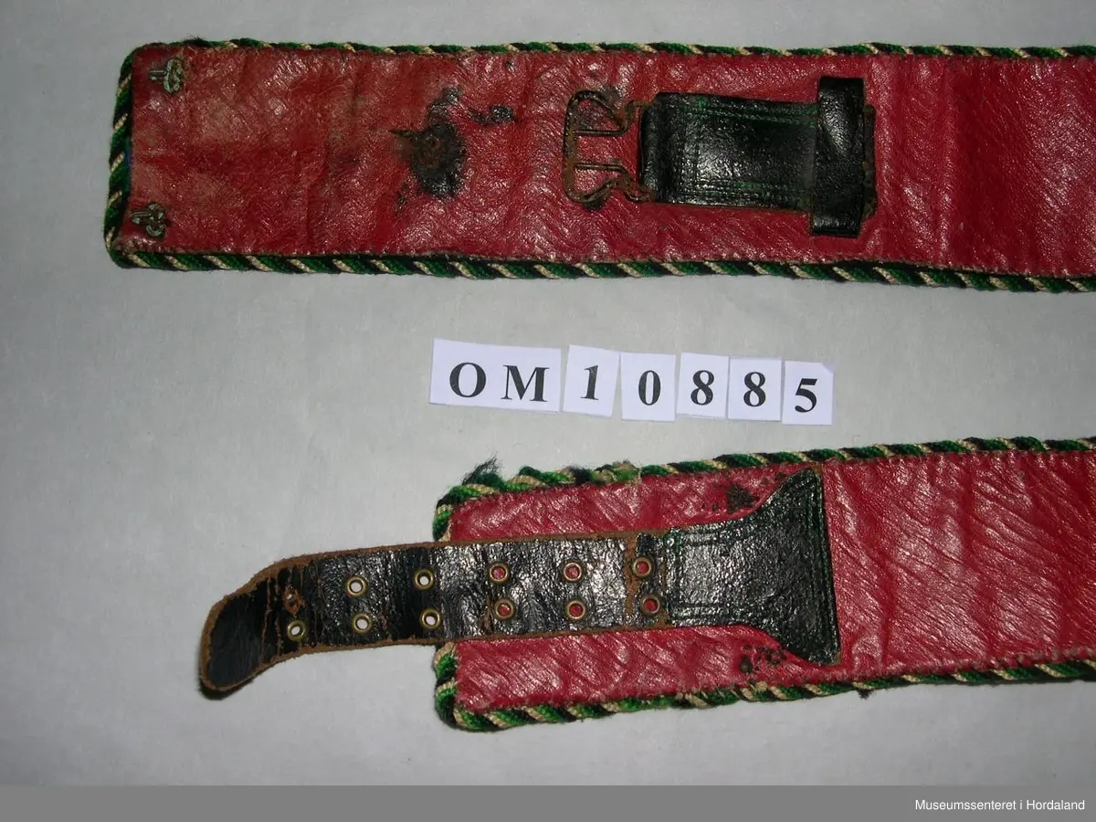 Brodert mannsbelte. Blomstermotiv med perledekor. Svart hovudfarge, blomster i grønt, gult, blått og oransje. Blanke og gullfarga småperler, nokre store, korte rørperler i gullfarge. I eine enden er det initialar og årstal, OASG 1887, sydde med perler som har rusta. Tvinna, fleirfarga snor til kanting. Raudt lêr til bakstoff. Beltet har beltespenne på baksida litt inn på beltet, og skinnreim med hol i andre enden. I tillegg er det fleire maljer og hekter.