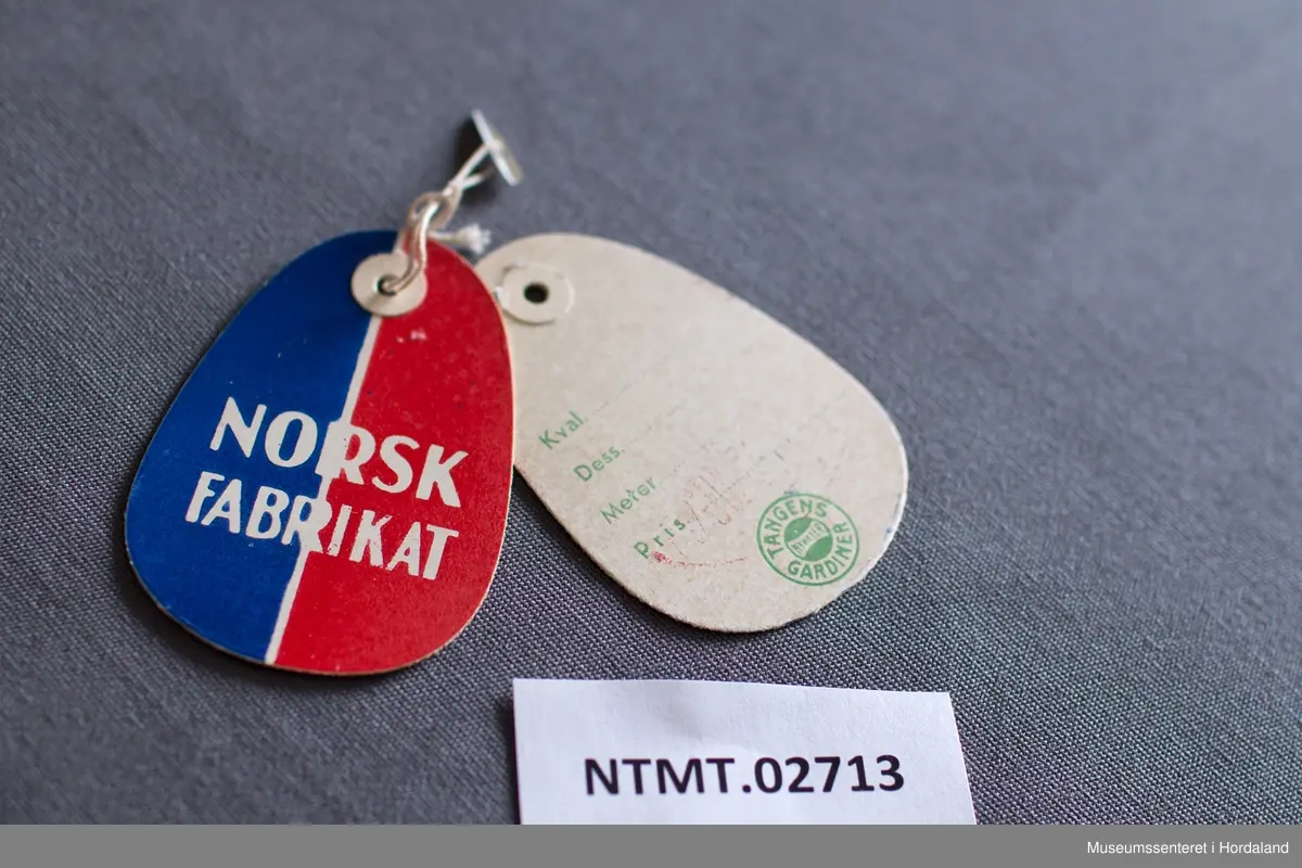 Raud og blå papiretikett/merkelapp frå Tangens gardiner.

Frå boka "Tangens Gardinfabrikk" (1950):
«Til å begynne med spant man ikke silke på å fremheve varens norske oprinnelse. Det var jo en tid da alt fint skulde være utenlandsk. Men fabrikken fastholdt prinsippet, og i det lange løp har den vunnet på det. Den beviste at norsk varer er kvalitetsproduksjon. Og nu vet alle at så er tilfelle.