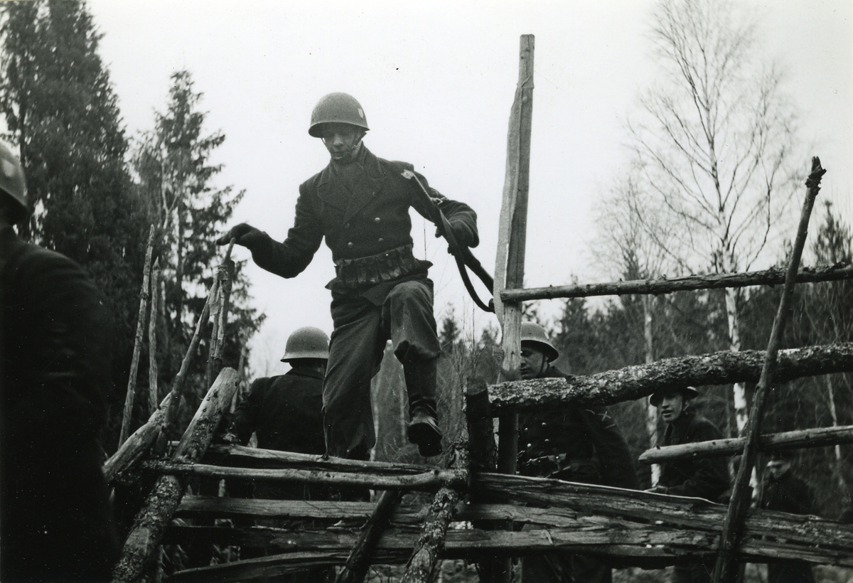 Manskap med hjälmar och karbiner tar sig över en gärdsgård.