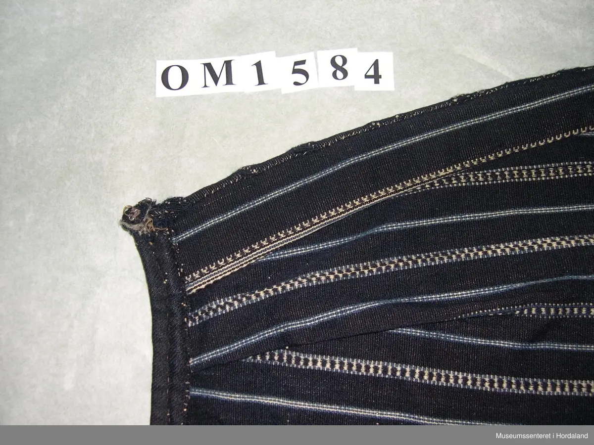 Mørkeblått, vove ullforkle med striper i kvitt og mellomblått. Linning. Glatt stykke midt framme, to folder på kvar side. Ei hekte til attlat.