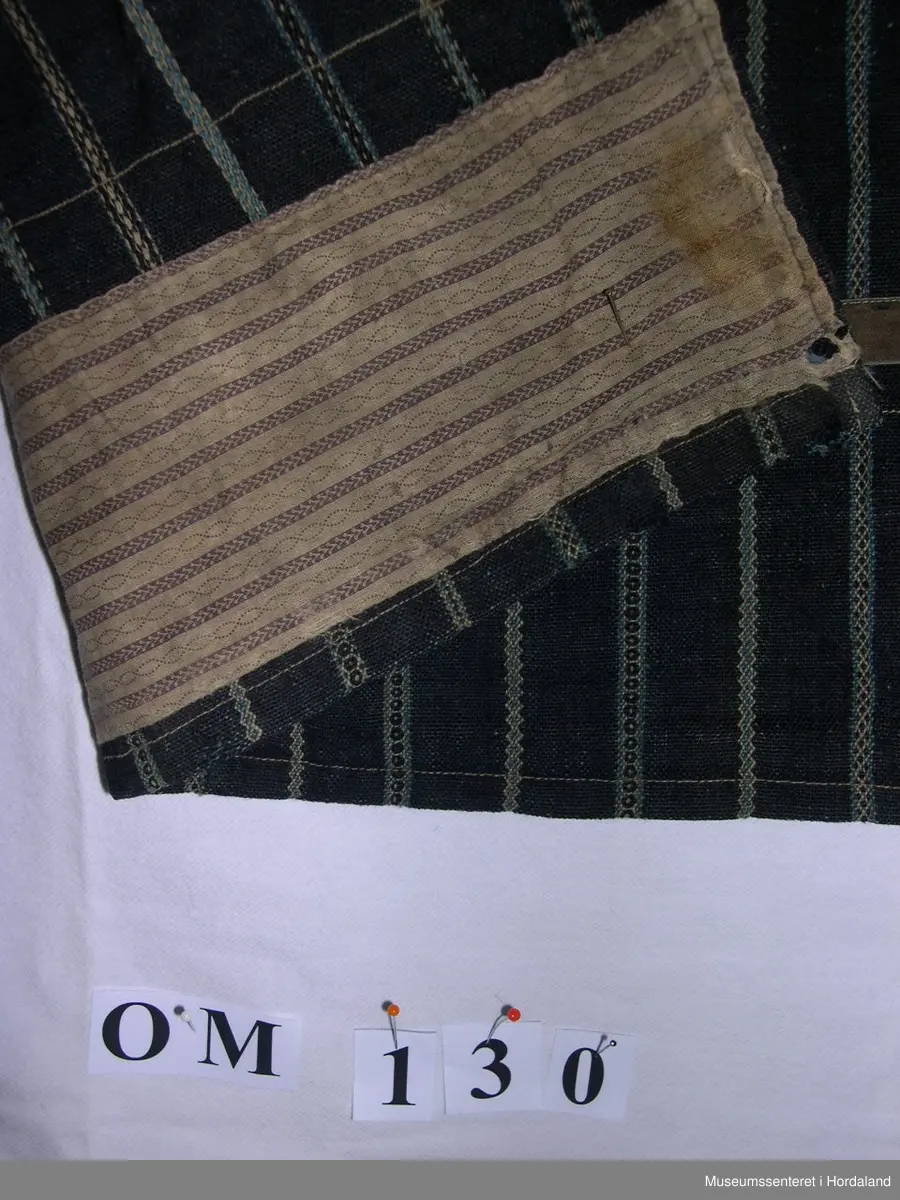 Mørkeblått, stripete vove forkle i ull. Striper i kvitt og blått mønster. Linning. Glatt stykke midt framme, tre folder på kvar side. Brunt fløyelsband nede. Skoning på baksiden med mønstra bomullsstoff og saum som syner på framsida. Forkle er smalt oppe. Holete.