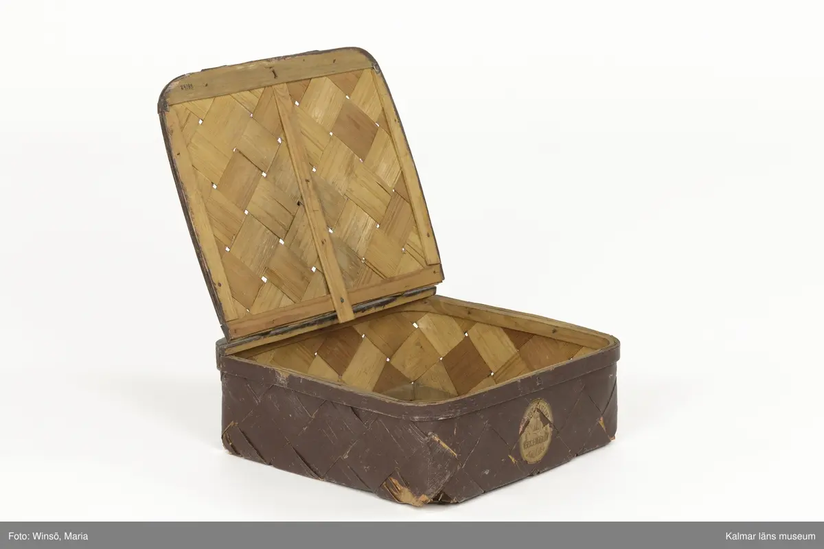 KLM 24189. Korg, konditorikorg. Fyrkantig, platt korg av flätat plattskuret spån. Korgen har ett lock med handtag och gångjärn av läder. Låsanordning av metall. Brunmålad med spår av grå färg på vissa ställen. På framsidan en tryckt etikett med text: August Sahlins Conditori Kalmar. Korgen är något trasig i botten.