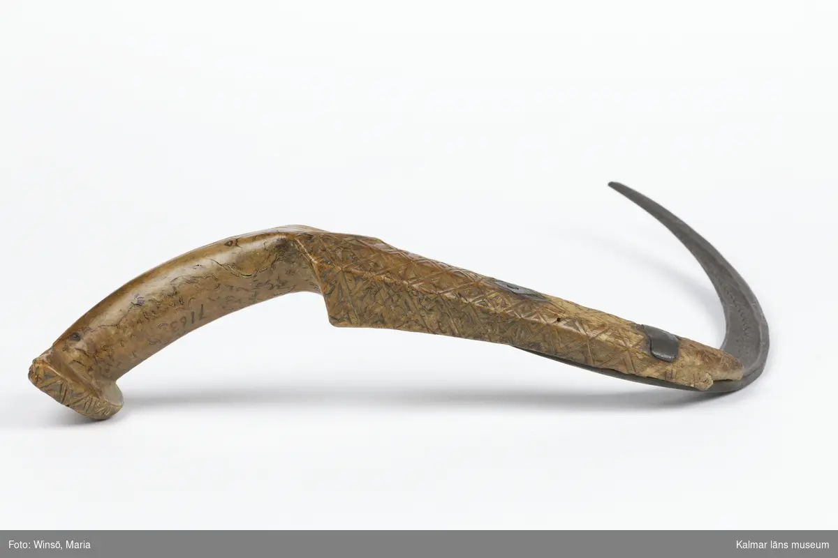 KLM 7163. Skära, vinkelskära. Av trä och metall. Vinkelskära med bågböjt blad av järn, fastnitat i skaftet. Skaft av masurbjörk, ornerat med plattsnidat rutmönster. Ornerat vågmönster på bladets ena sida samt inpunsat: ANO 1788 SIS. Rund knopp, en bit saknas. På knoppen skuret sicksackmönster samt 1788 SIS. Användes för att skära av säden.