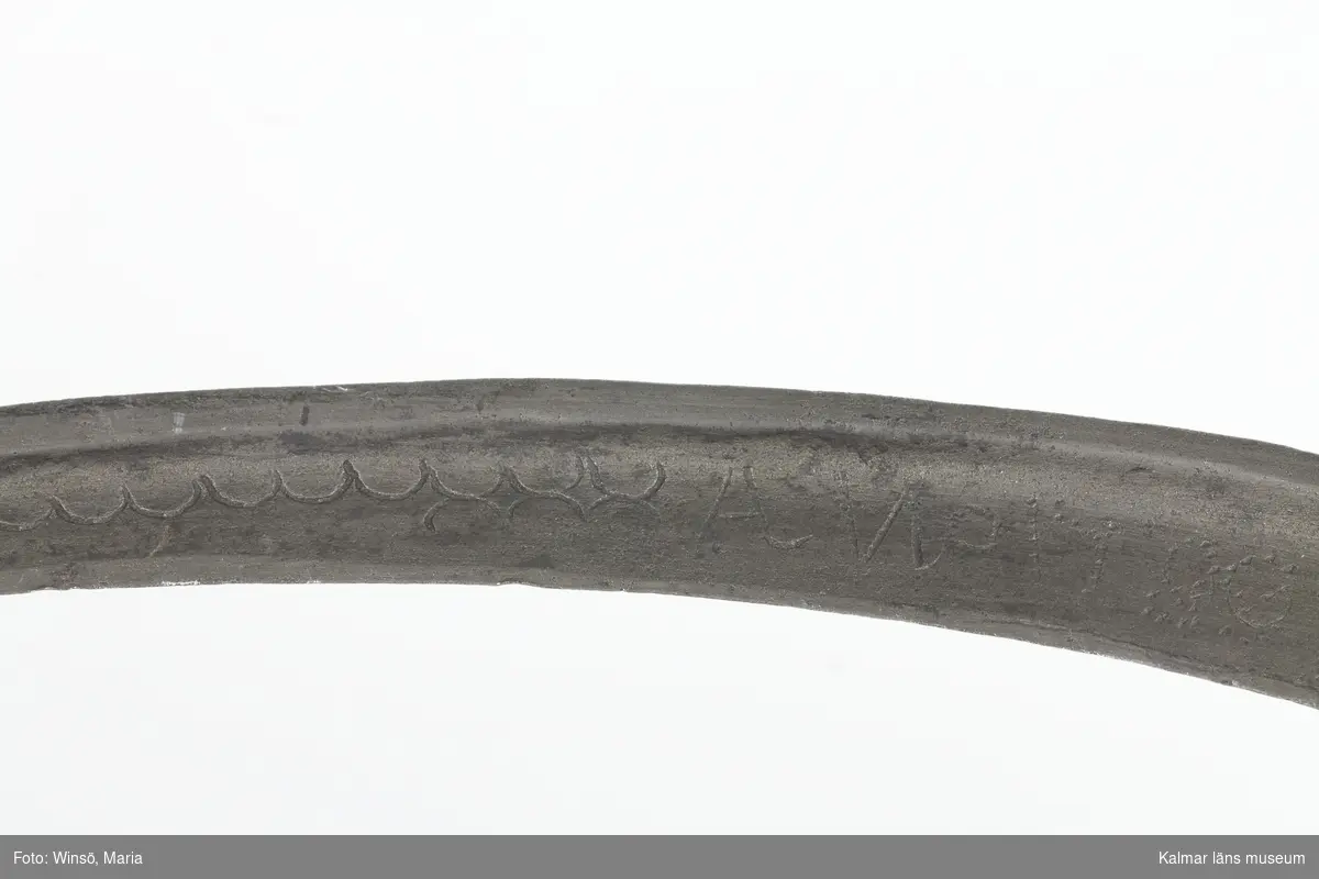 KLM 7163. Skära, vinkelskära. Av trä och metall. Vinkelskära med bågböjt blad av järn, fastnitat i skaftet. Skaft av masurbjörk, ornerat med plattsnidat rutmönster. Ornerat vågmönster på bladets ena sida samt inpunsat: ANO 1788 SIS. Rund knopp, en bit saknas. På knoppen skuret sicksackmönster samt 1788 SIS. Användes för att skära av säden.