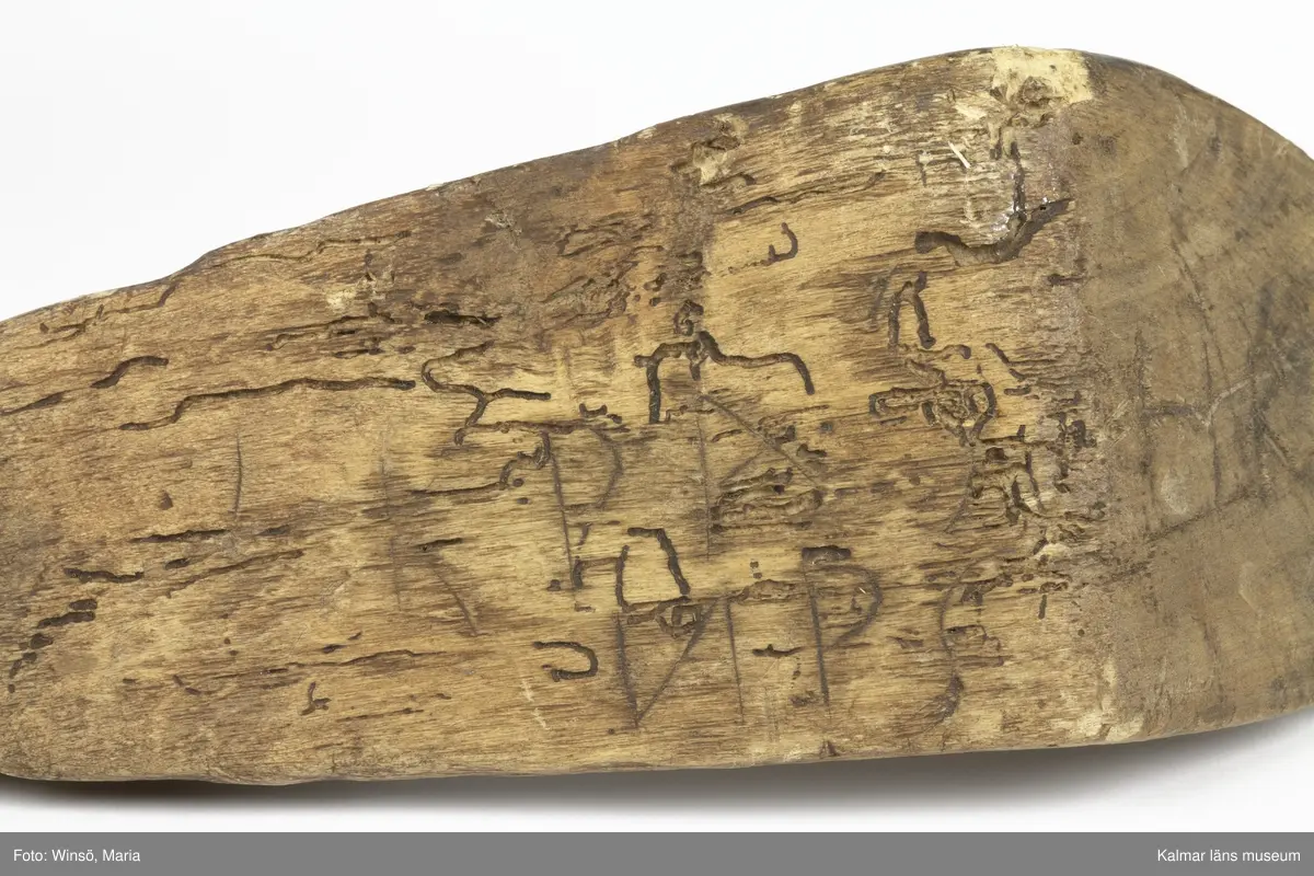 KLM 15141. Skära, vinkelskära, trä, metall. Vinkelskära med bågböjt blad av järn, fastnitat på skaftets ena sida. Skaft av trä med handbygel. Bladet är målat i rödbrun färg, för rostskydd, under senare tid. Inristning skaft: KPA, NPS HR. Stämpel blad: lis ?