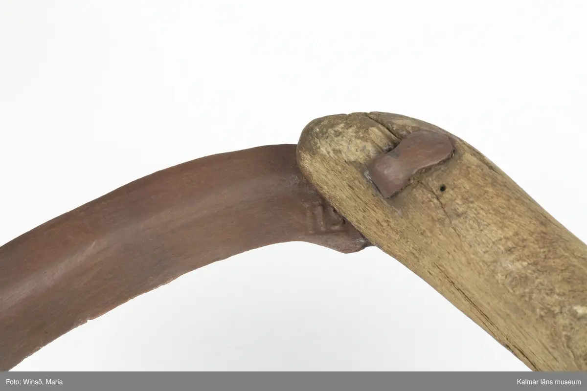 KLM 15141. Skära, vinkelskära, trä, metall. Vinkelskära med bågböjt blad av järn, fastnitat på skaftets ena sida. Skaft av trä med handbygel. Bladet är målat i rödbrun färg, för rostskydd, under senare tid. Inristning skaft: KPA, NPS HR. Stämpel blad: lis ?