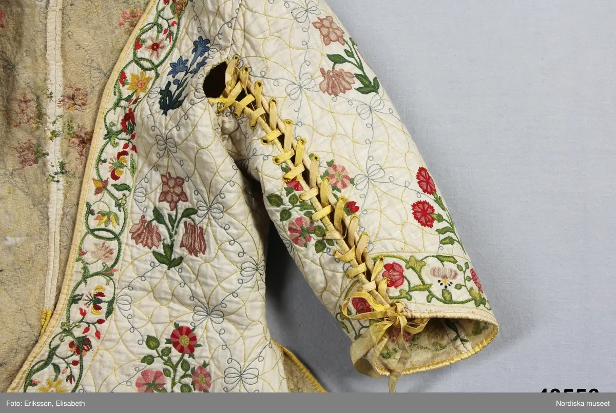 Katalogkort IV:
"Tröja / kofta, för dam, 1600-talets slut.
Längd 54 cm
Av linnelärt med foder av glest tuskaftat ylletyg. Försedd med stickningar i gult och blått samt broderier i kedje-söm med yllegarn. 2 ryggstycken, 2 framstycken. Halvlånga ärmar med 2 sömmar, varav den ena öppen och kantad med gult siden samt försedd med gult silke tränsade snörhål, med band av dubbelvikt sammansytt band. Sömarna uppslitsade nedifrån upptill midjan och kantade med gult siden."