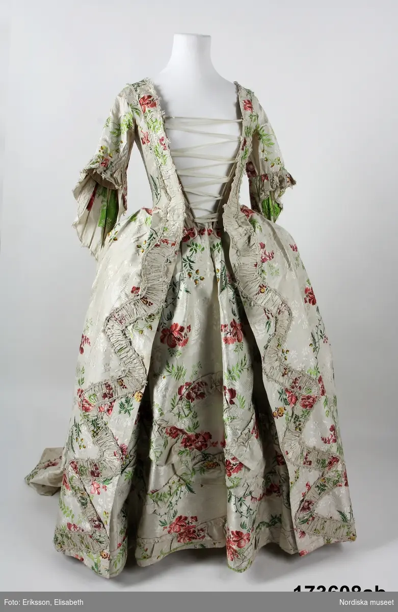 Äldre katalogkort: 
"Robe och kjol  av vit sidenbrokad med blommor i rött, gult m.m. Robe med dubbla watteauveck och engageanter av vita knypplade silkespetsar i mångdubbla rader. Livet fodrat med vitt linne med fiskben i sömmarna. Bröstlapp saknas.
Tygets vådbredd 50,5 cm. Tygåtgång ca 13,5 meter."

Bandgarnering av veckad remsa av tyget kantat med uddganser i vitt silke.
I utmärkt fint skick.
/Berit Eldvik 2009-01-09