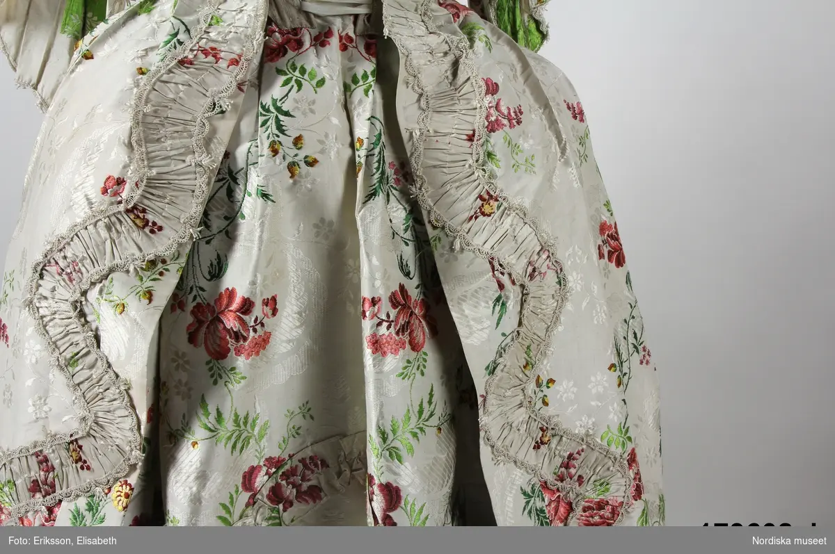 Äldre katalogkort: 
"Robe och kjol  av vit sidenbrokad med blommor i rött, gult m.m. Robe med dubbla watteauveck och engageanter av vita knypplade silkespetsar i mångdubbla rader. Livet fodrat med vitt linne med fiskben i sömmarna. Bröstlapp saknas.
Tygets vådbredd 50,5 cm. Tygåtgång ca 13,5 meter."

Bandgarnering av veckad remsa av tyget kantat med uddganser i vitt silke.
I utmärkt fint skick.
/Berit Eldvik 2009-01-09