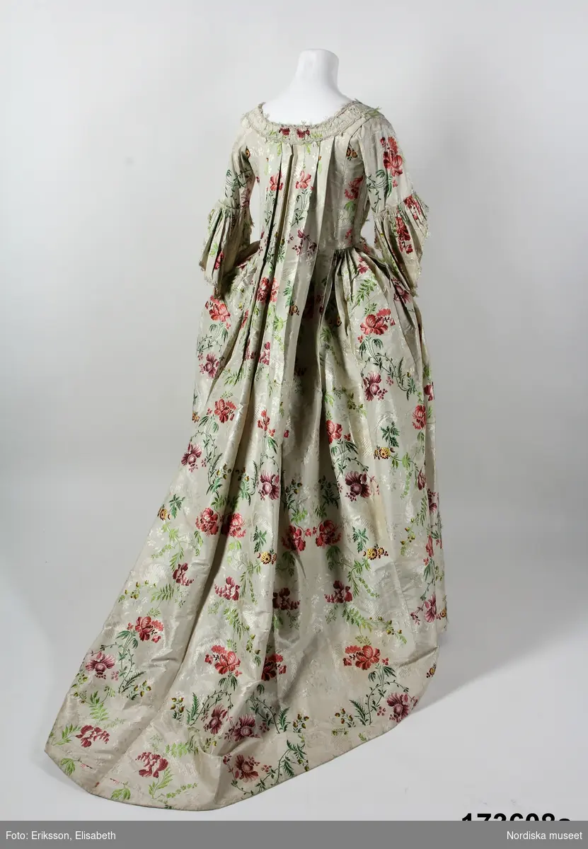 Äldre katalogkort: 
"Robe och kjol  av vit sidenbrokad med blommor i rött, gult m.m. Robe med dubbla watteauveck och engageanter av vita knypplade silkespetsar i mångdubbla rader. Livet fodrat med vitt linne med fiskben i sömmarna. Bröstlapp saknas.
Tygets vådbredd 50,5 cm. Tygåtgång ca 13,5 meter."

Bandgarnering av veckad remsa av tyget kantat med uddganser i vitt silke.
I utmärkt fint skick.
/Berit Eldvik 2009-01-09