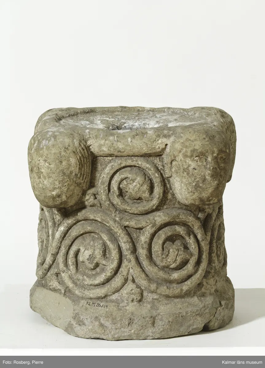 KLM 35314. Dopfunt, dopfuntsfot. Av kalksten. Fot till dopfunt med rund genom skärning. Spiralupprullade växtrankor i relief samt upptill fyra kraftigt utstående ansiktsmasker. Ett cylindrisk hål tvärs igenom stenen.