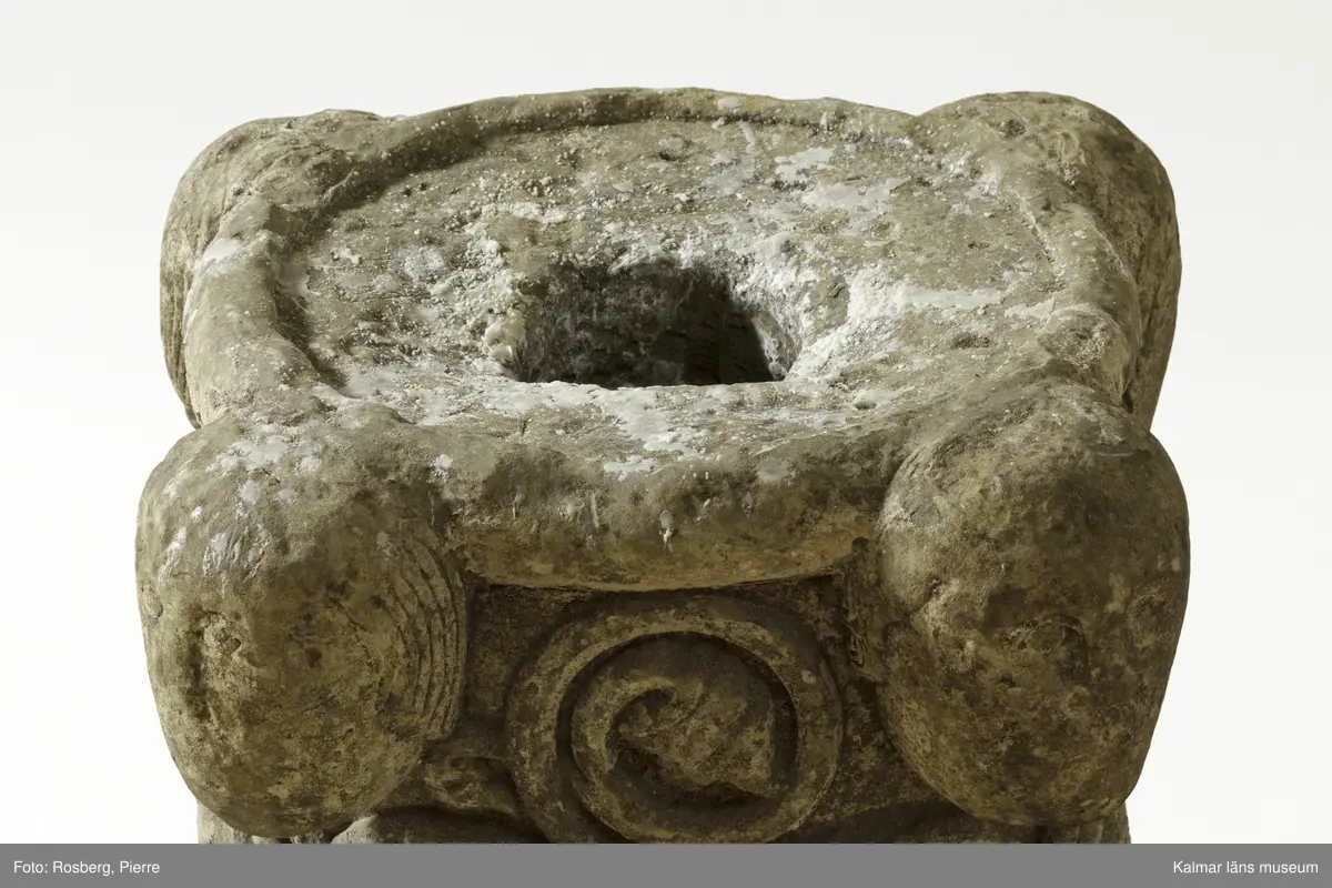 KLM 35314. Dopfunt, dopfuntsfot. Av kalksten. Fot till dopfunt med rund genom skärning. Spiralupprullade växtrankor i relief samt upptill fyra kraftigt utstående ansiktsmasker. Ett cylindrisk hål tvärs igenom stenen.